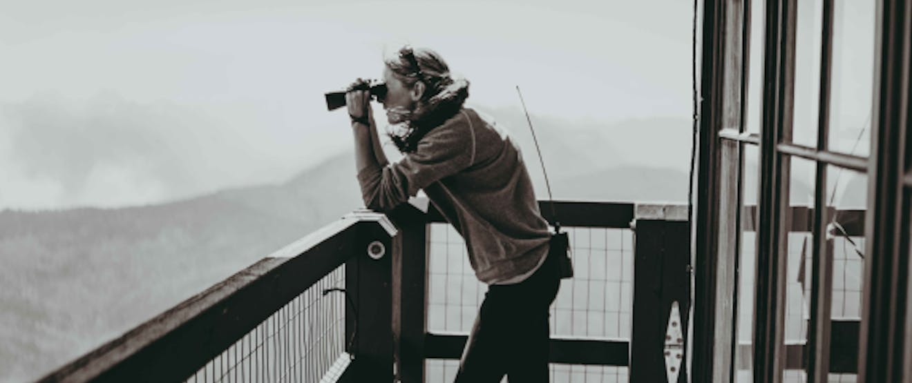 black and white of a woman on the deck of a lookout towner looking out at something through binoculars