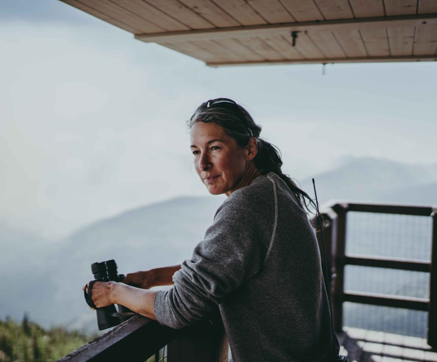 a close up image of a woman wearing grey sweatshirt, holding binoculars standing on the catwalk of a fire lookout tower