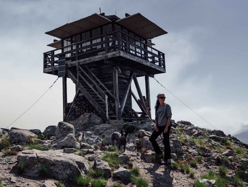 a woman wearing a grey sweatshirt and black pants standing next to her black and grey cattle dog on the of a rocky peak where a fire lookout stands