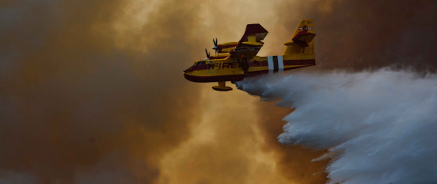 an areal photo of a yellow and red forest service plane dropping water on a raging fire above the treelike