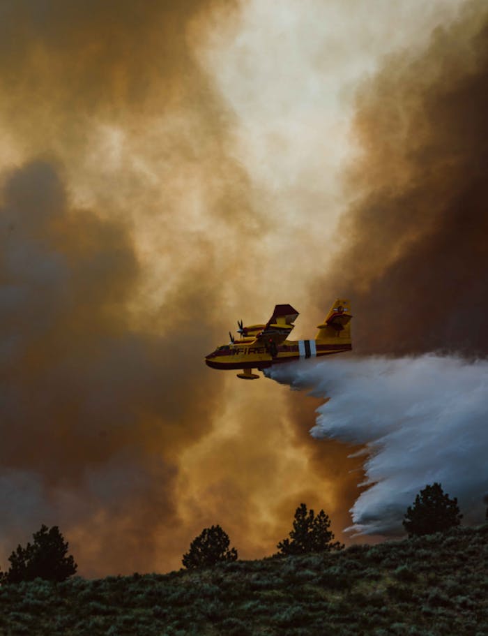 an areal photo of a yellow and red forest service plane dropping water on a raging fire above the treelike