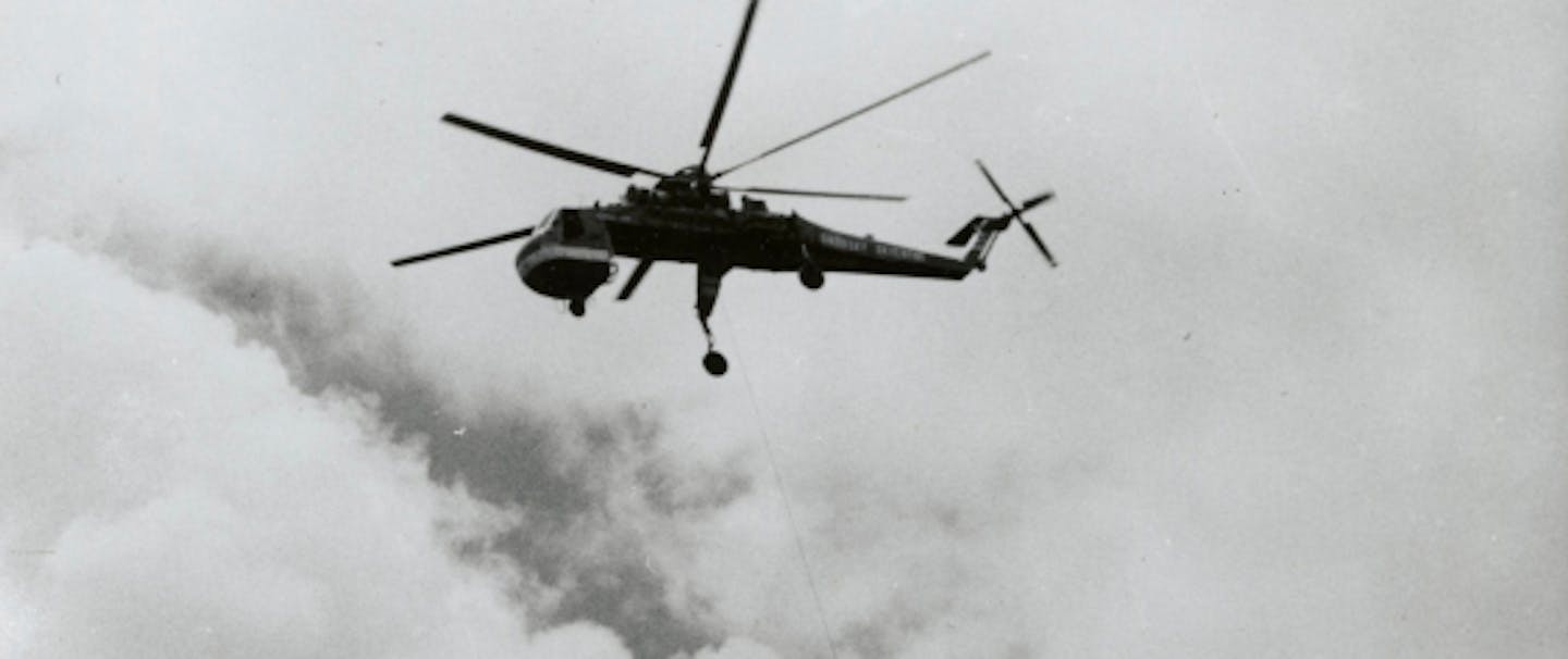 a black and white image of a helicopter flying up to pull the hanging log to the drop zone away from the logging site