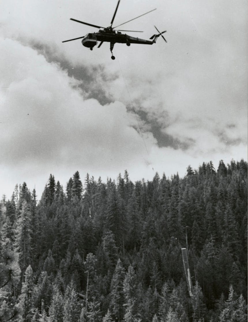 a black and white image of a helicopter flying up to pull the hanging log to the drop zone away from the logging site