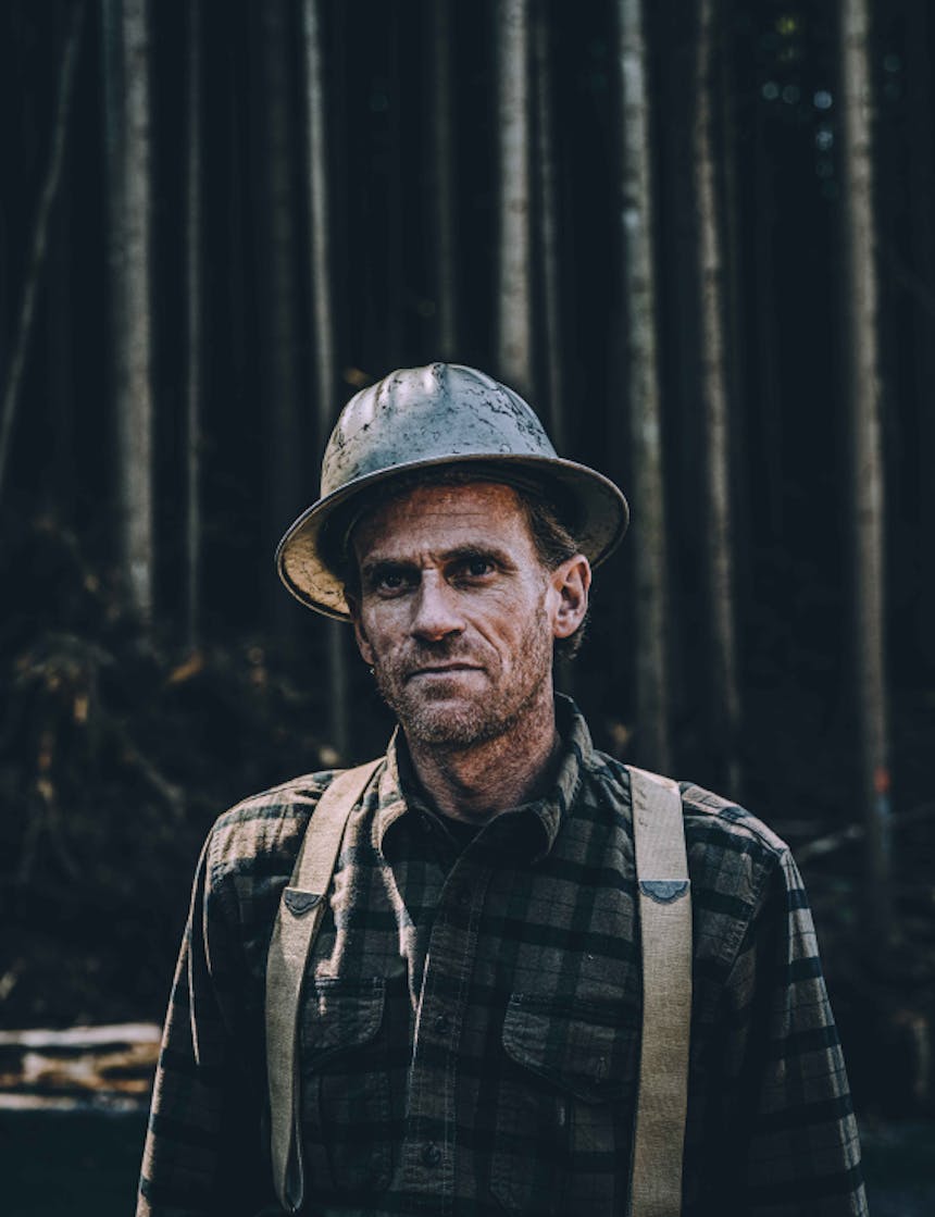 a portrait of a scruffy bearded logger wearing a grey hardhat, brown plaid flannel and brown suspenders at a logging operation