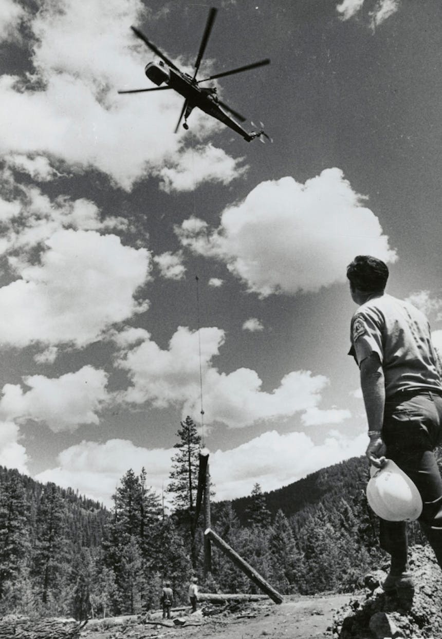 black and white image of a USFS man in uniform holding a white hardhat, looking up to a helicopter pulling a couple of freshly cut logs away as two men stand near those logs further ahead