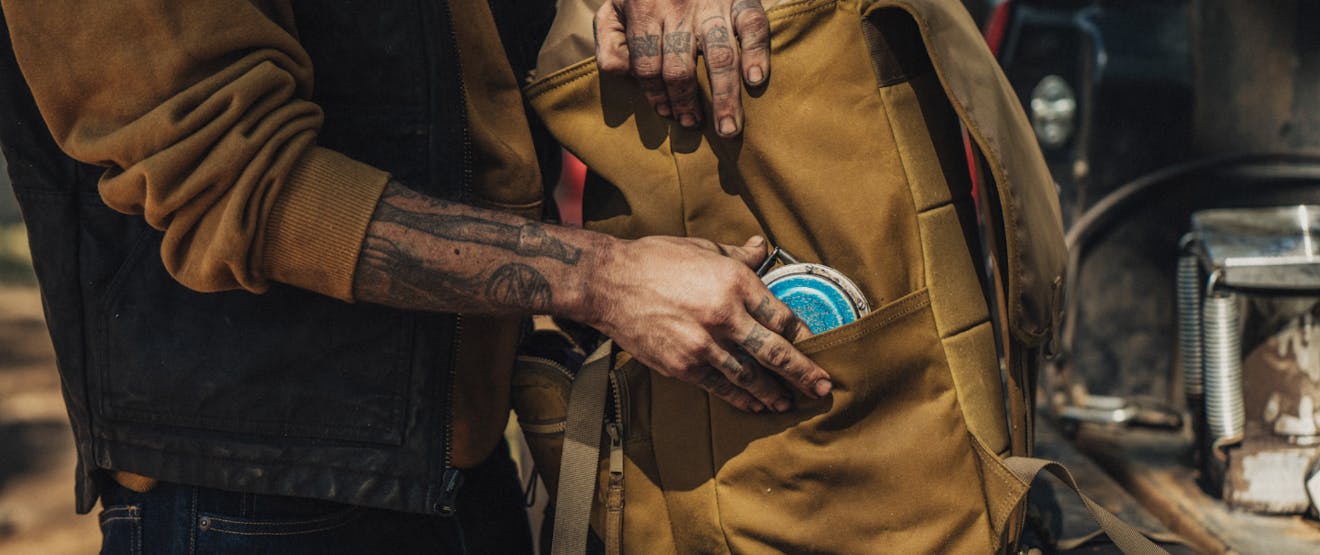 close up image of tattoos on a mans arms and hands, wearing a light brown sweatshirt under and black work vest sliding a blue measuring tape into the side pocket of a similarly brown colored backpack