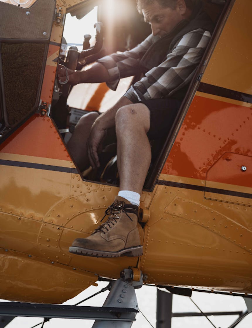 View from the side of a yellow and orange float plane with a middle aged white man wearing a grey flannel and work boots working with gears
