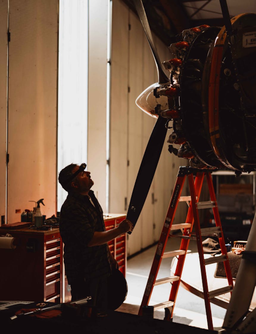 a white man wearing a headlamp looking at the propeller of a float plane in a workshop