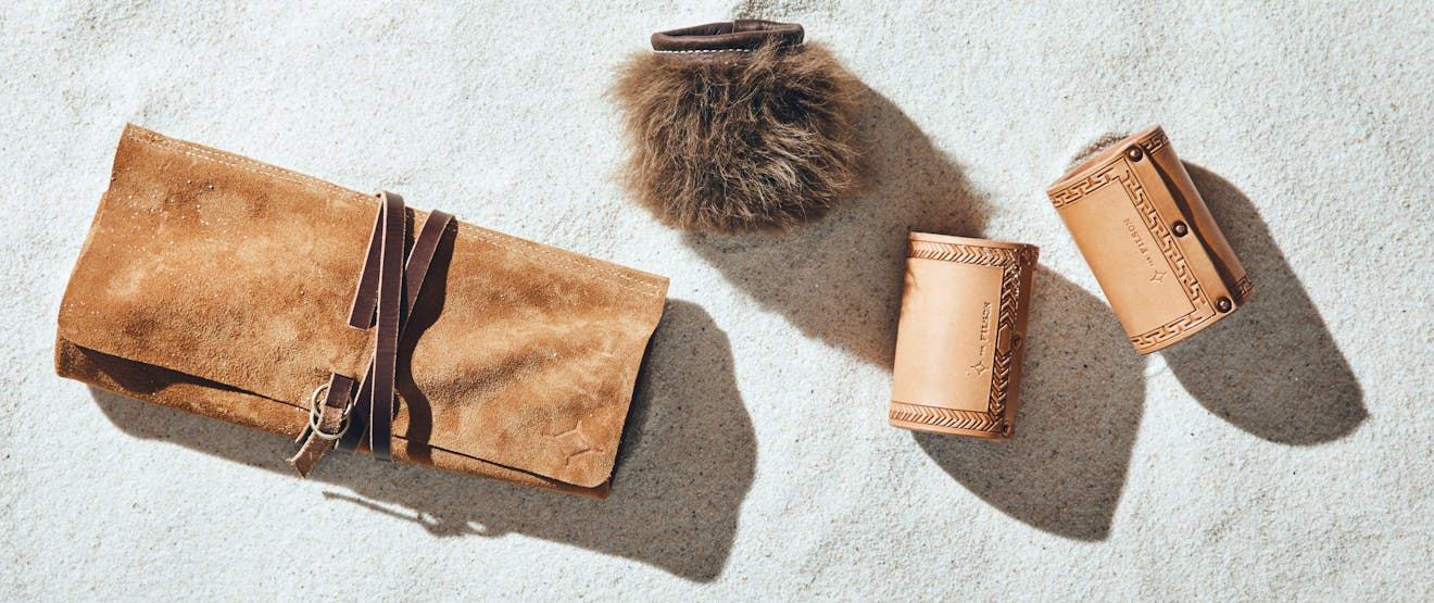 four products laid in white sand, a bison fur coozie, two leather coozie, and cow hide tool roll.