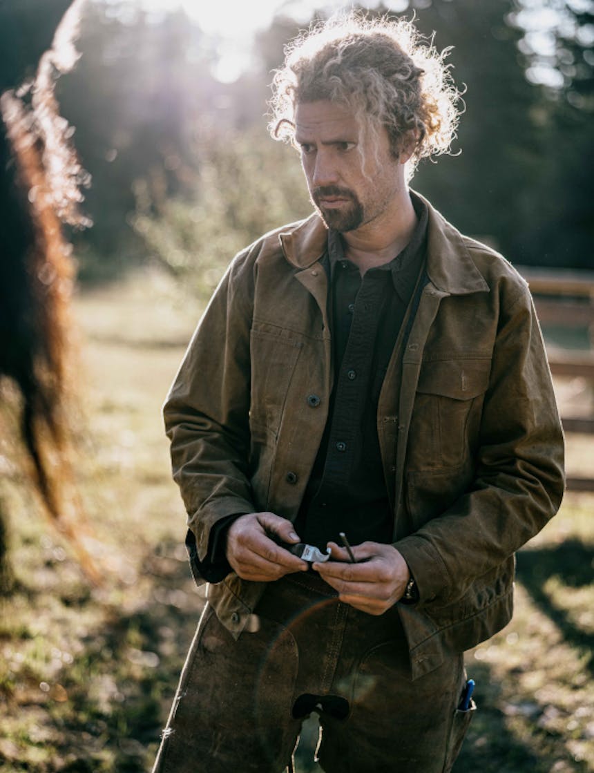 a wavy blonde haired man wearing workwear clothes looking at his horse