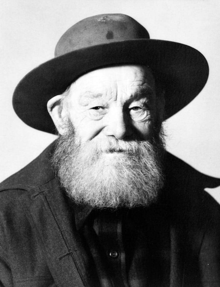 a black and white portrait of an old bearded wrinkly man wearing a wool brimmed hat, wool coat and flannel shirt