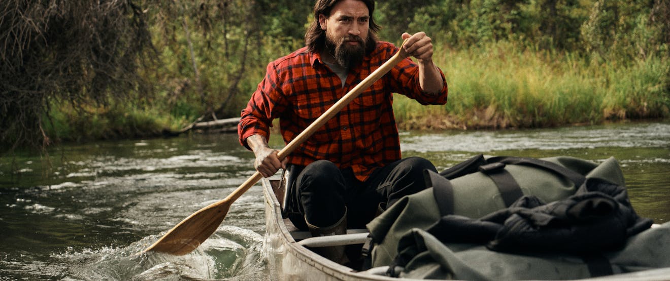 a native man wearing a red and black flannel and black pants, paddling a canoe through a river filled with green dry bags