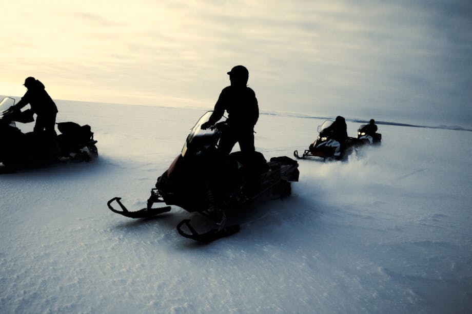 four people on snow machines in the early light of day driving across an ice sheet in Alaska