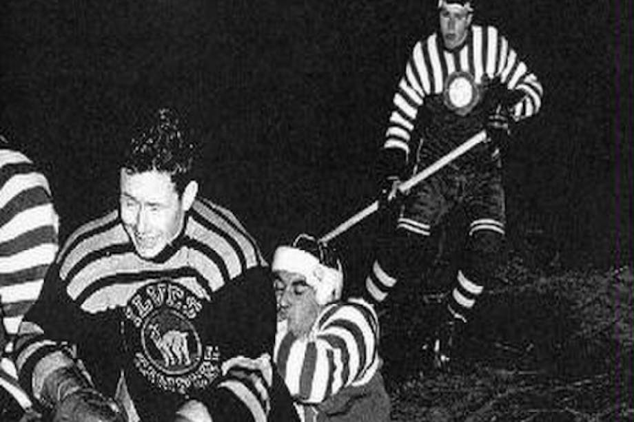 black and white historic photo of hockey players colliding mid ice, one falling behind the other