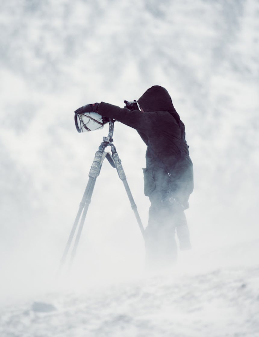 man wearing full clothing shooting with a large tripod and camera lens as the winds blow snow around him