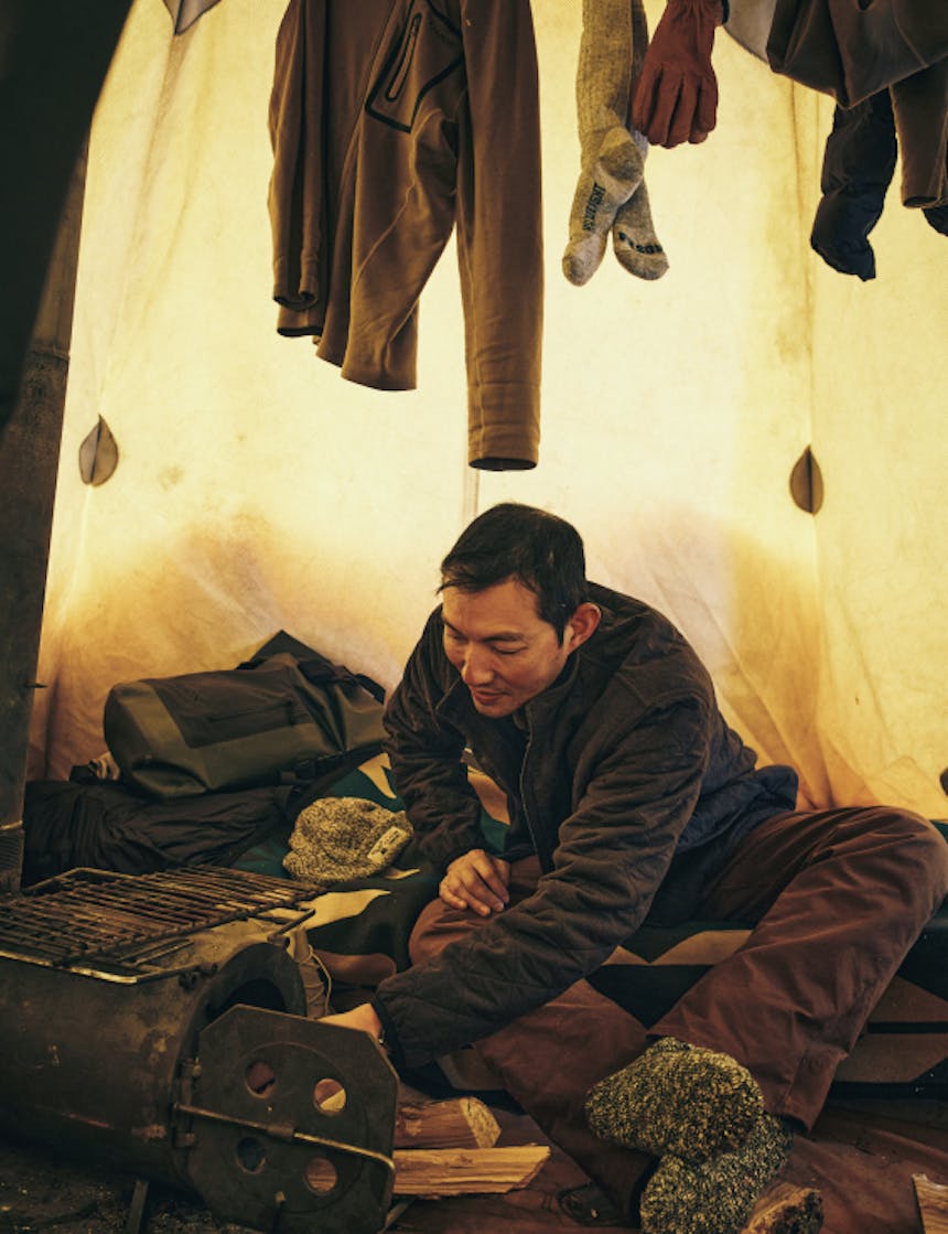 an indigenous man inside a tent feeding the wood stove as clothes hang above to dry