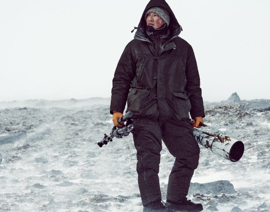 an indigenous man wearing full layers holding camera gear in either hand standing on rocky arctic tundra