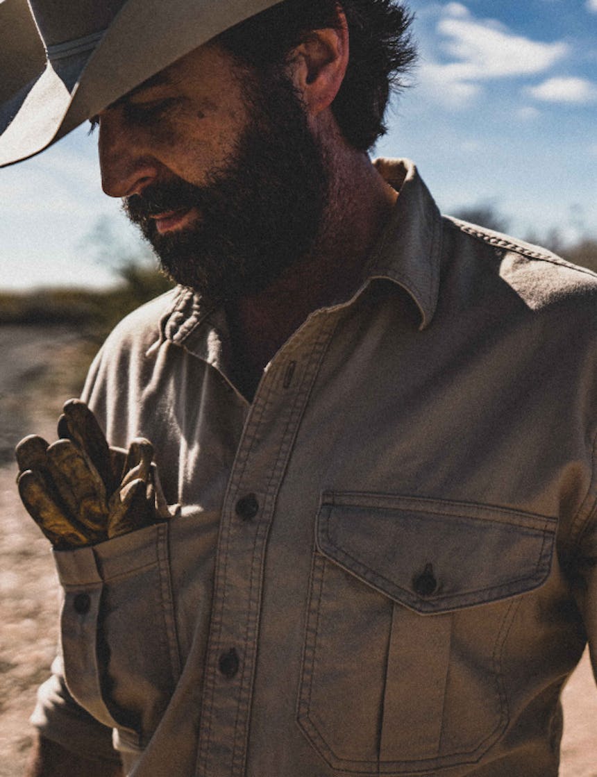 close up of a bearded man wearing a cowboy hat and tan button up with gloves tucked into his top right pocket