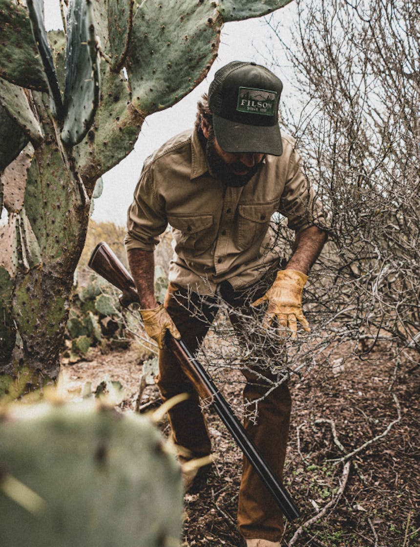 a man wearing a green baseball cap, tan button up, yellow gloves and brown pants moving between brush and a cactus