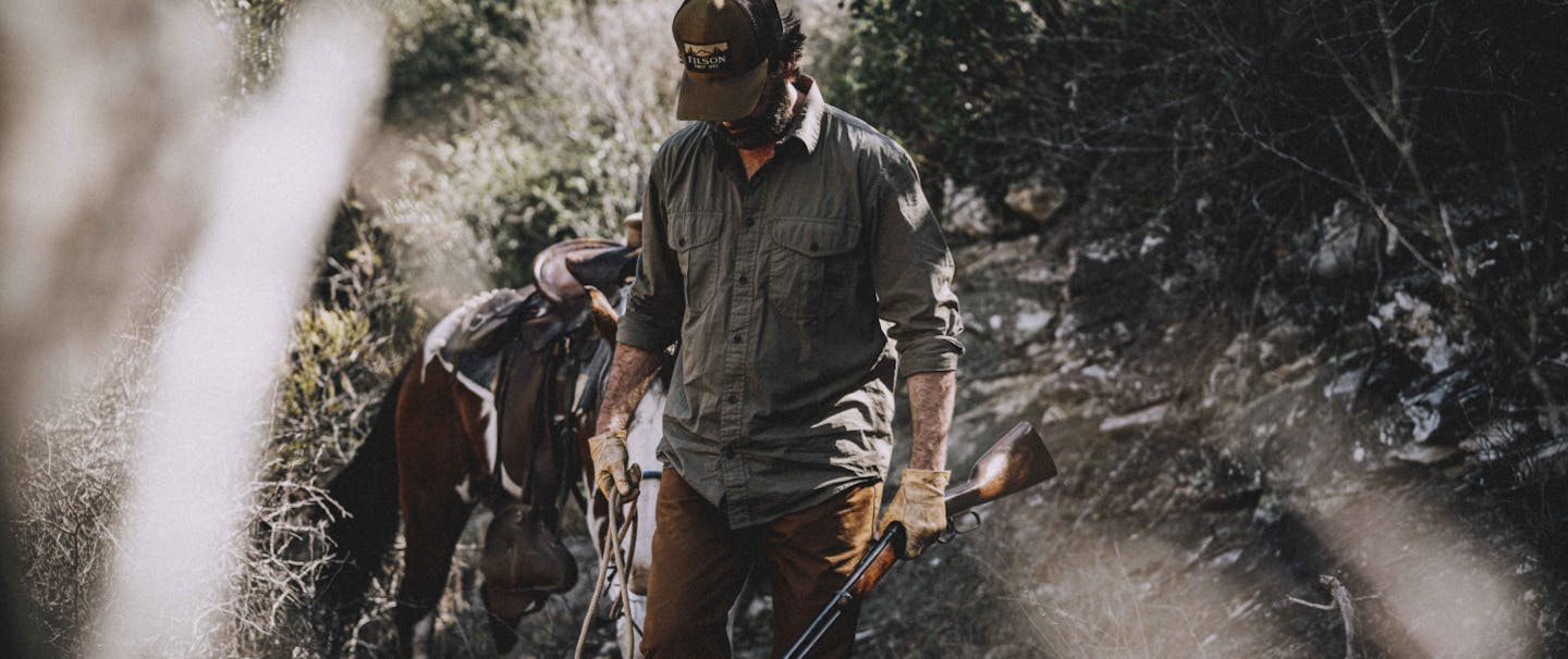 bearded man wearing a brown baseball cap, grey button up and brown pants walking through brush with a horse holding a gun