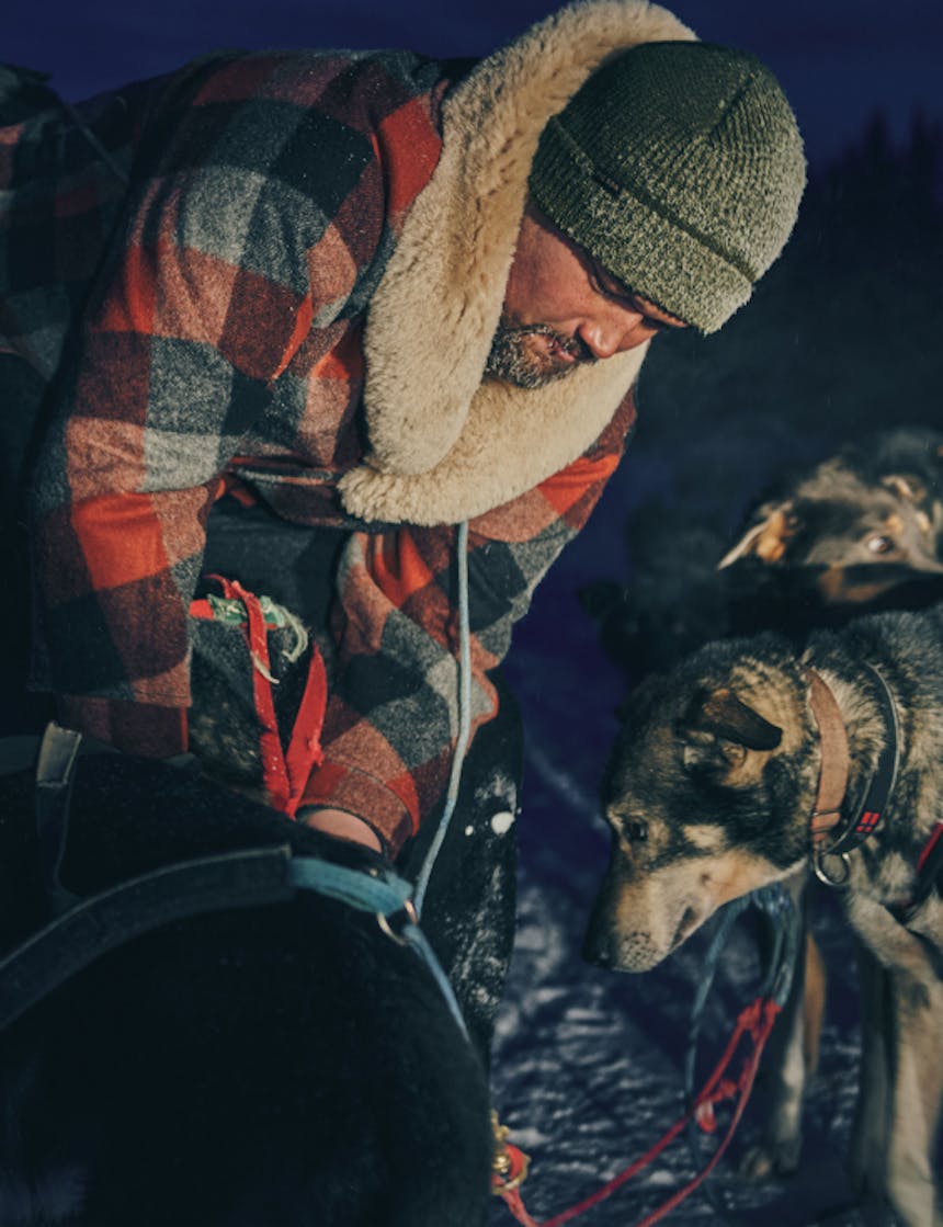 a close up of a man tending to one of his sled dogs wearing a grey beanie and red, black and grey wool coat