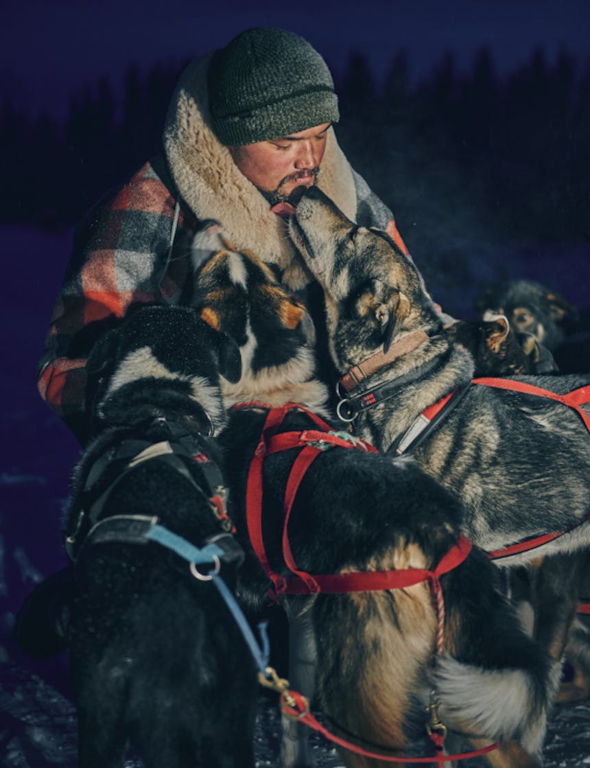 close up of a musher petting three of his dogs, one licking his face, in at lantern light as it gets darker out around them