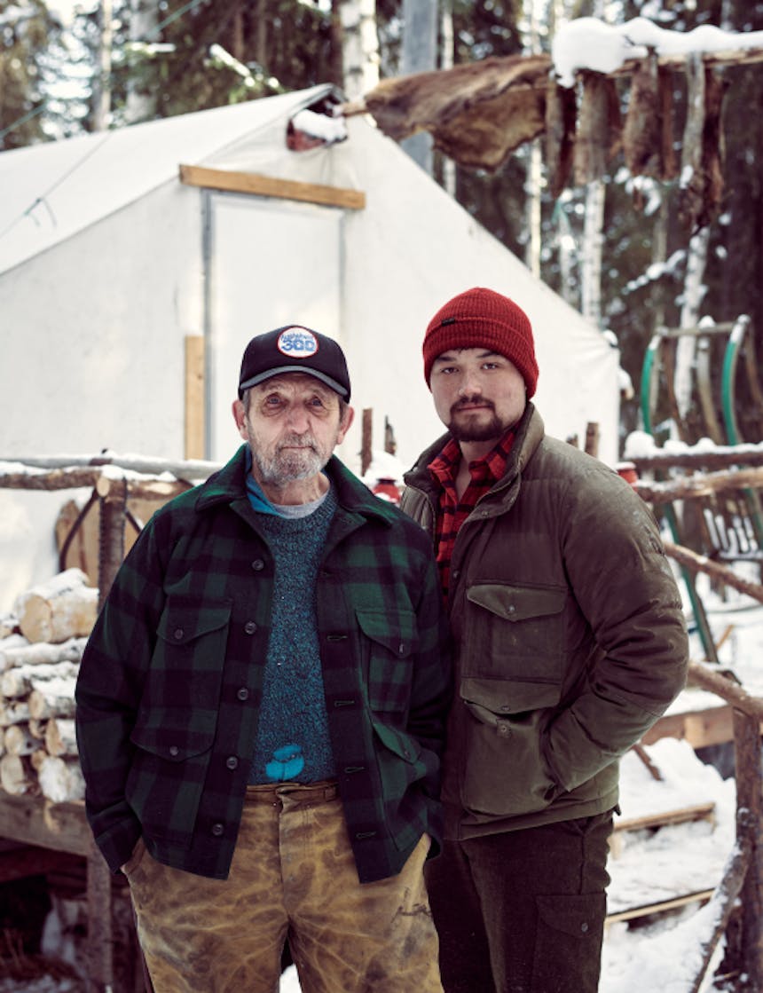 a father son photo standing outside of their wall tent, the father on the left old and wrinkled wearing a black baseball cap, grey sweater and green and black plaid wool jacket; the son on the left wearing a red beanie, brown down jacket over a red and black flannel