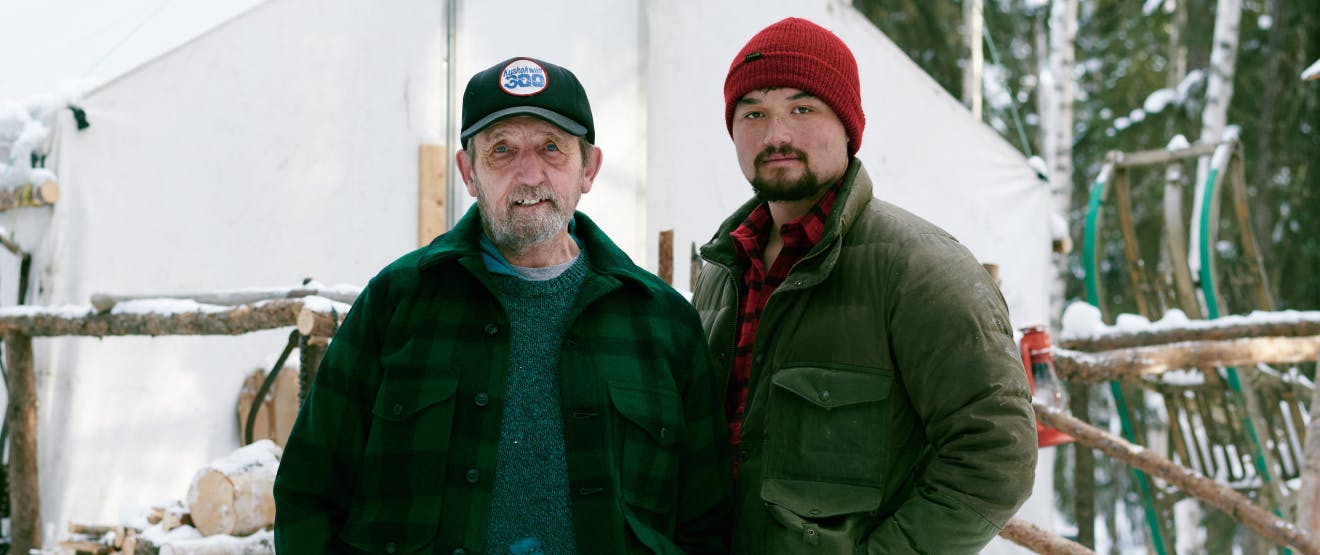 a father son photo, the father on the left old and wrinkled wearing a black baseball cap, grey sweater and green and black plaid wool jacket; the son on the left wearing a red beanie, brown down jacket over a red and black flannel