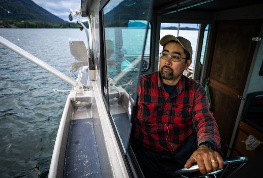 a close up of a man wearing glasses and a red and black plaid shirt driving a fishing boat as nets are strung off the back