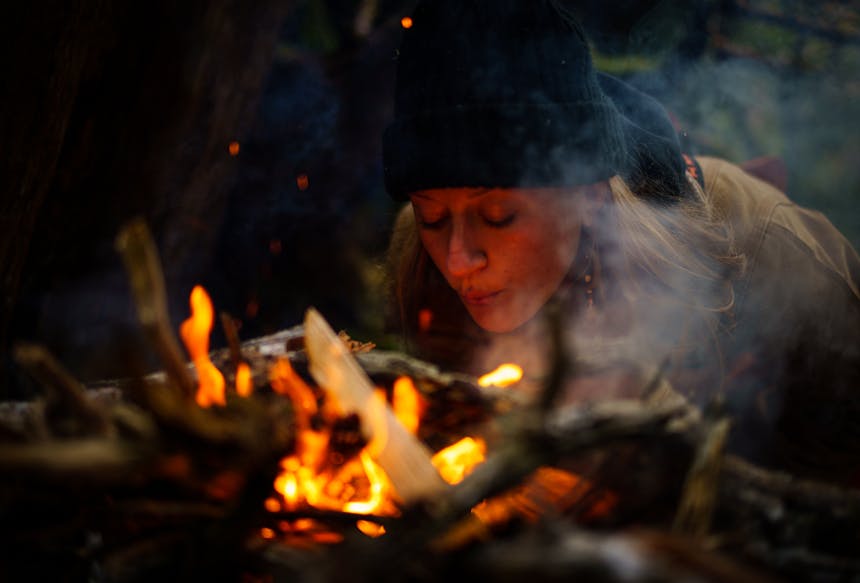 a close up of a blonde woman wearing a black knit cap and brown jacket blowing on a fire to get it started