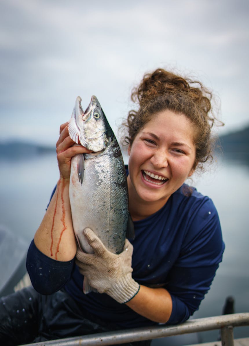 a smiling an happy brown haired native alaskan woman, holding a freshly caught fish as blood drips down her arm