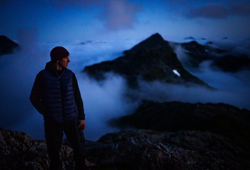 a man wearing a knit cap and down jacket looking off to the right during blue hour on a mountain top with peaks in the background