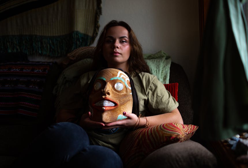 a woman sitting in a living room chair, pillows, blankets and curtains around her as she holds a native mask proudly