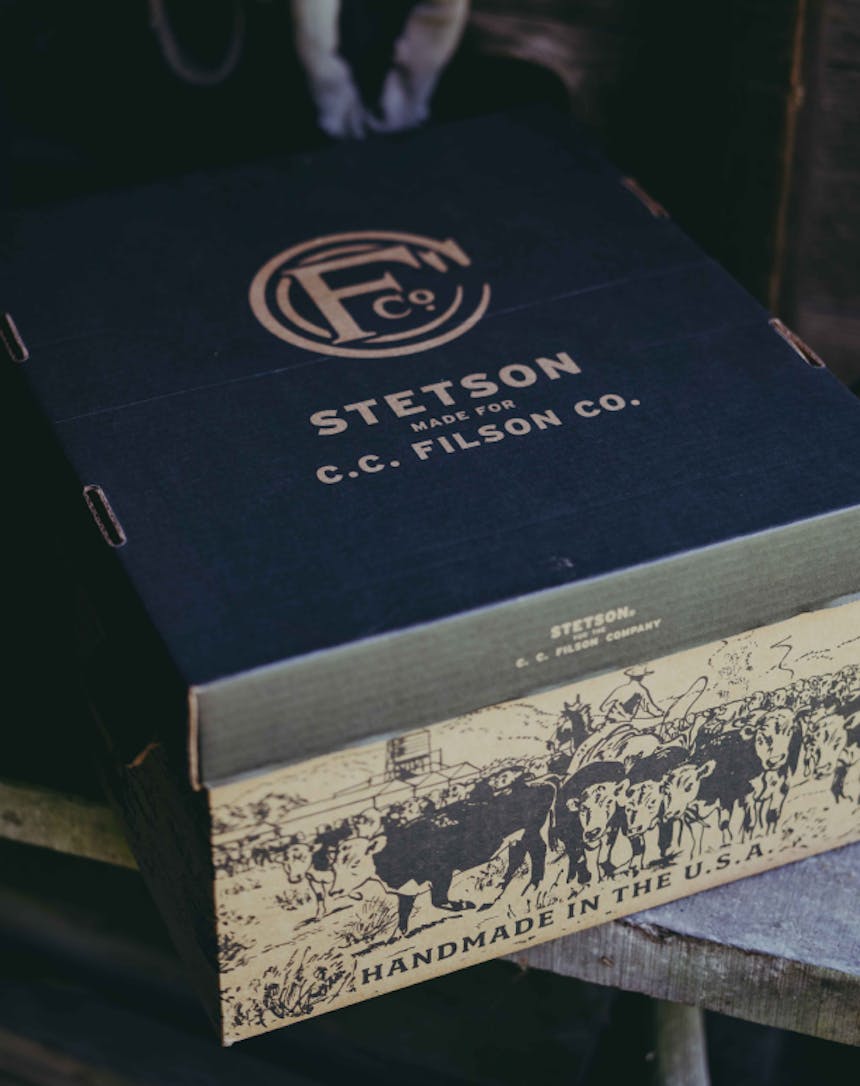 a cardboard box sitting on a wooden bench in a barn, the box reading Stetson Made for CC Filson Co