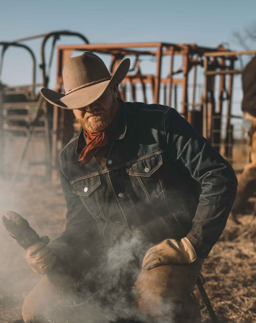 a close up of a cowboy wearing a denim jacket holding a cows leg while it gets branded