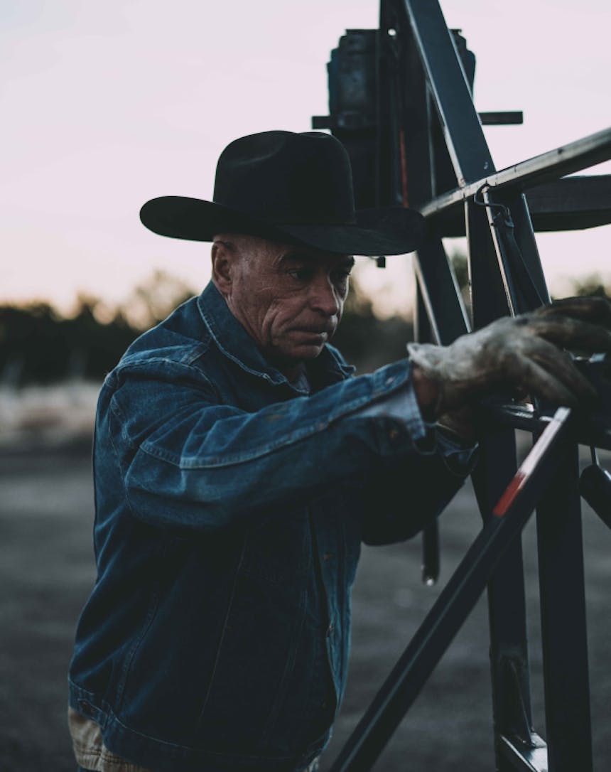 an older cowboy wearing a denim shirt and black cowboy hat adjusting the settings on a cattle shoot as the sun sets