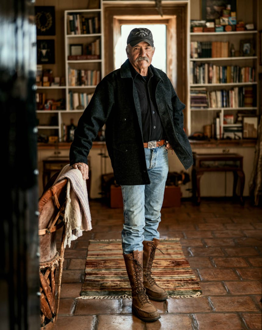 grey haired white man standing Lin an office wearing a baseball cap, black button up shirt, grey wool jacket, jean and tall leather boots