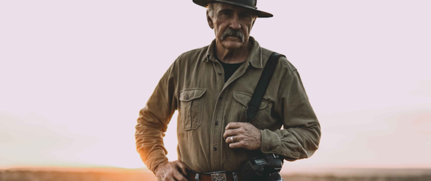 grey haired white man wearing a cowboy hat, brown button up shirt, jean and tall leather boots holding a canon camera looking directly at the camera as he sun sets behind him