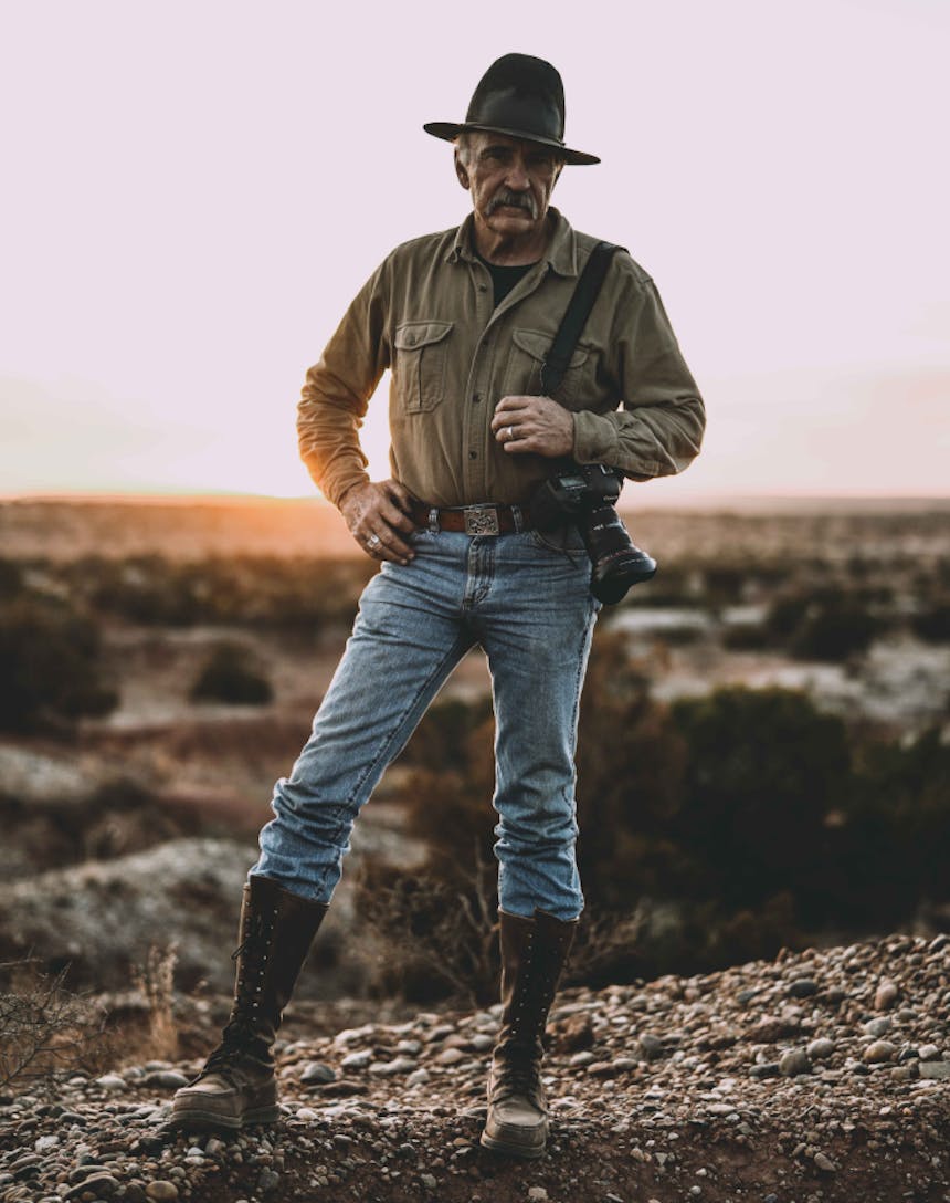 grey haired white man wearing a cowboy hat, brown button up shirt, jean and tall leather boots holding a canon camera looking directly at the camera as he sun sets behind him