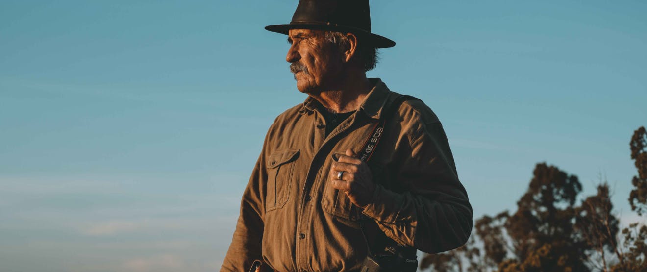 grey haired white man standing looking off to the sunset with a beautiful light blue light behind him, wearing a cowboy hat, brown button up shirt, jean and tall leather boots holding a canon camera