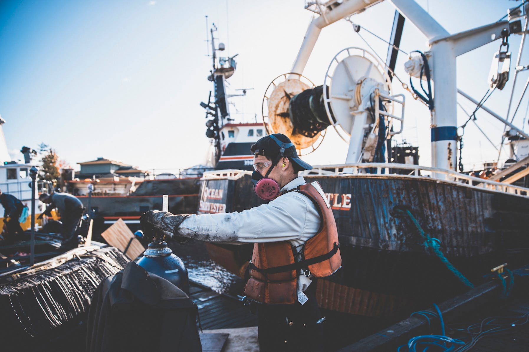 Caucasian man wearing a respirator and protective eyewear inspects a gas cylinder in the shipyard. Rugged fishing vessels with signs of rust docked in the background.