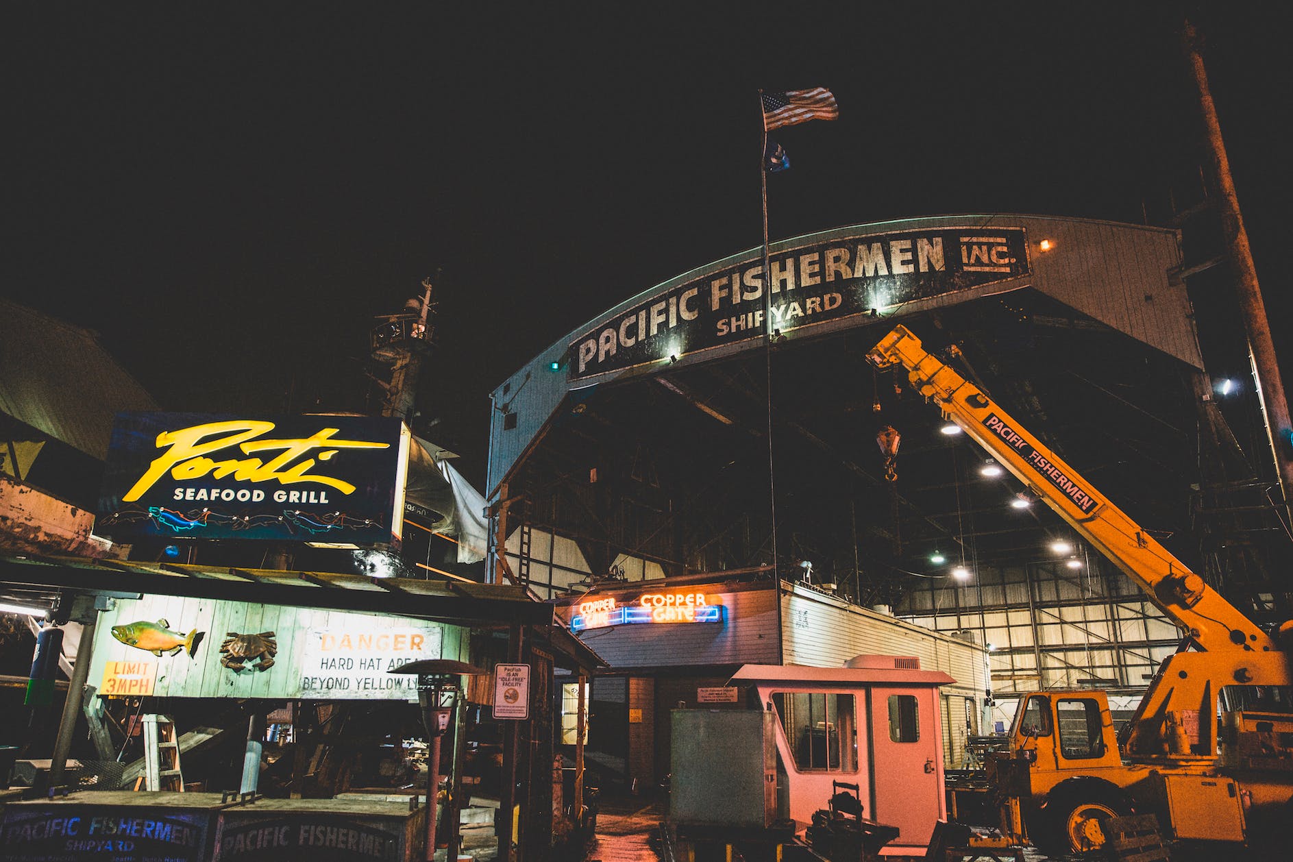Wide angle image of the Pacific Fishermen Shipyard at night, with neon signs and spotlights illuminating the structures. An American flag at full mast sits on a flag pole in the background.