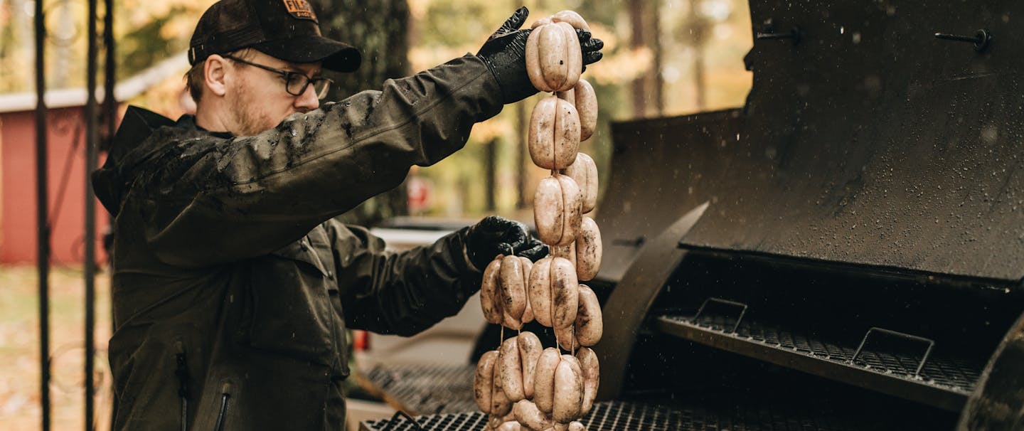 man in black coat sets linked sausages on large black grill outside in woods