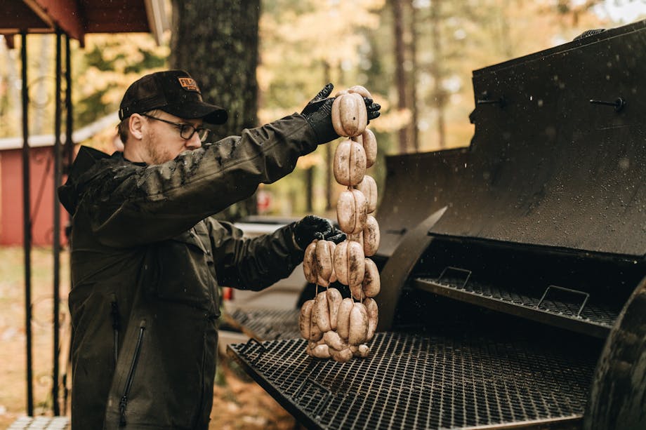 man in black coat sets linked sausages on large black grill outside in woods