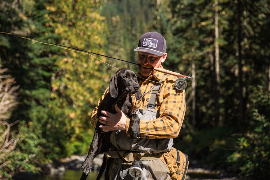 Forest setting. Man in a yellow flannel shirt holding a black dog with both hands, and a fishing rod clenched between his teeth.