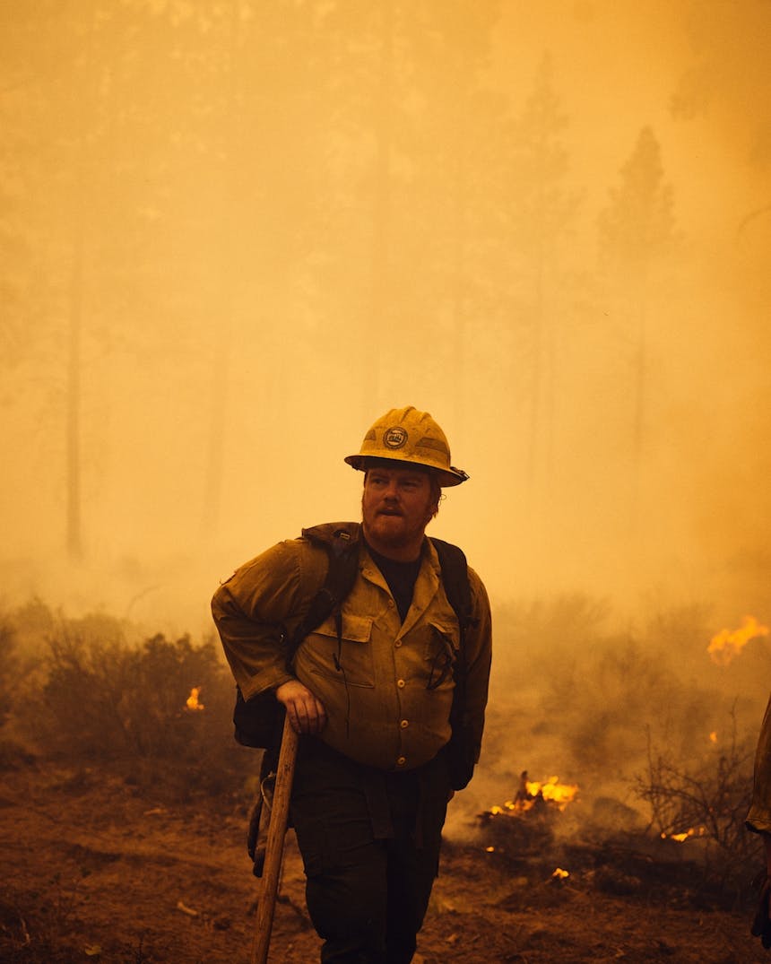 fire fighter in hard hat stands in smoky flaming field with hand on shovel