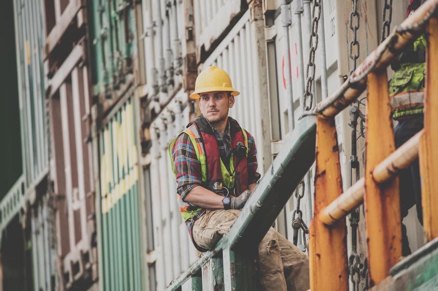 man in hard hat and safety vest sits on railing next to stacked shipping containers