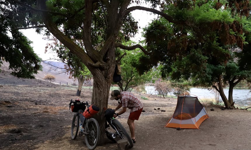 man working on bike resting on tree at campsite near river