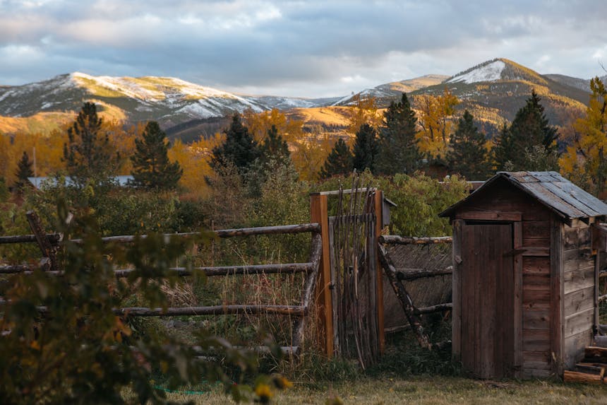 wooden outhouse in yard with autumnal forest and mountain backdrop