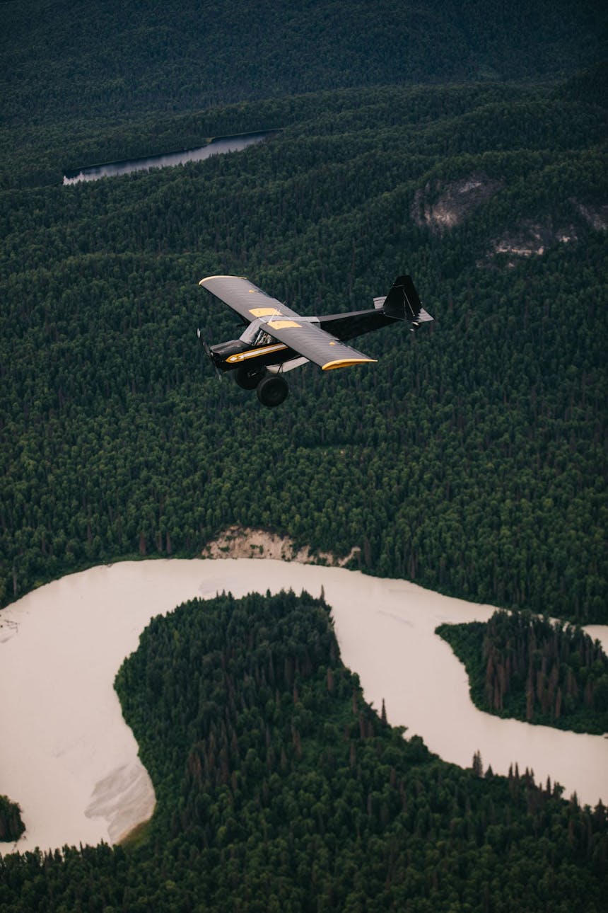 Sarah flying small black plane over river cutting through pine forest